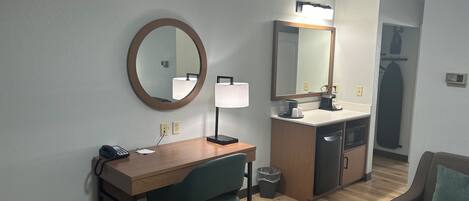 A hotel room with a wooden desk, a green office chair, a round mirror, a vanity with a mirror, a sink, a coffee maker, and a trash can.