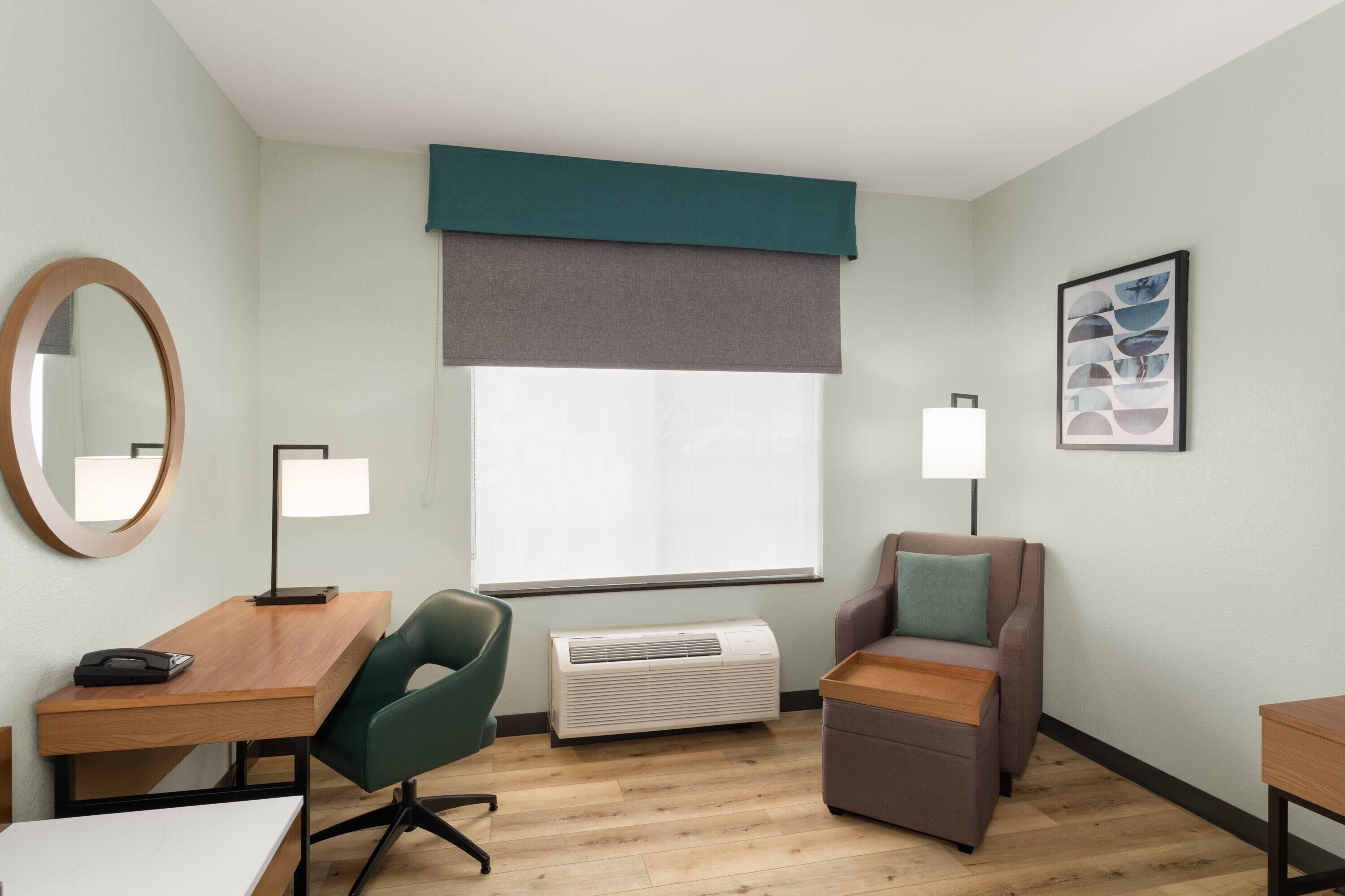 Habitación de hotel con un escritorio de madera, una silla verde, un sofá marrón, una lámpara de pie, un ventanal con vistas y un cuadro en la pared.