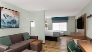 A hotel room with a sofa, a small table, a bed with a blue headboard, a TV mounted on the wall, and a window with a view. - Wingate Suites By Wyndham Mansfield (Mansfield)