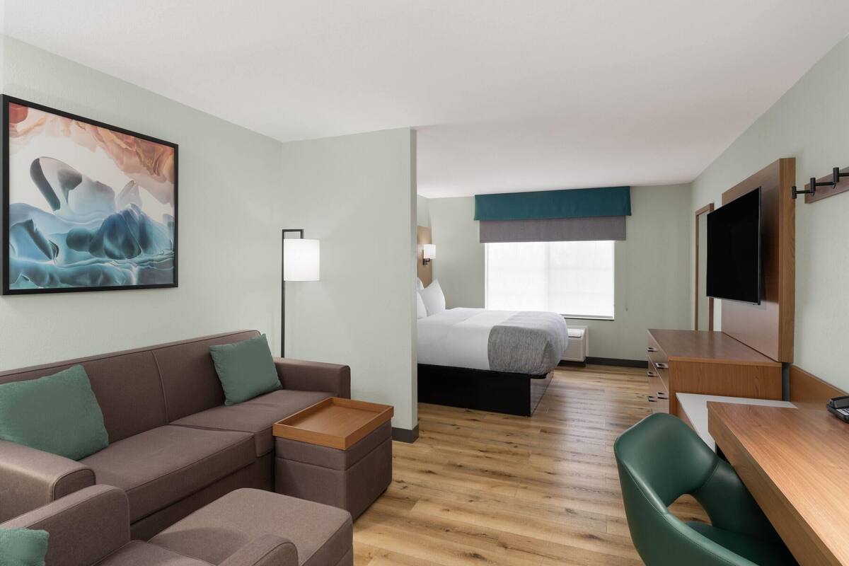 Ein Hotelzimmer mit einer Couch, einem kleinen Tisch, einem Bett mit blauer Kopfteil sowie einem an der Wand montierten Fernseher und einem Fenster mit Blick.