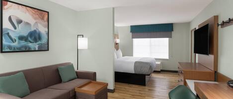 A hotel room with a sofa, a small table, a bed with a blue headboard, a TV mounted on the wall, and a window with a view.