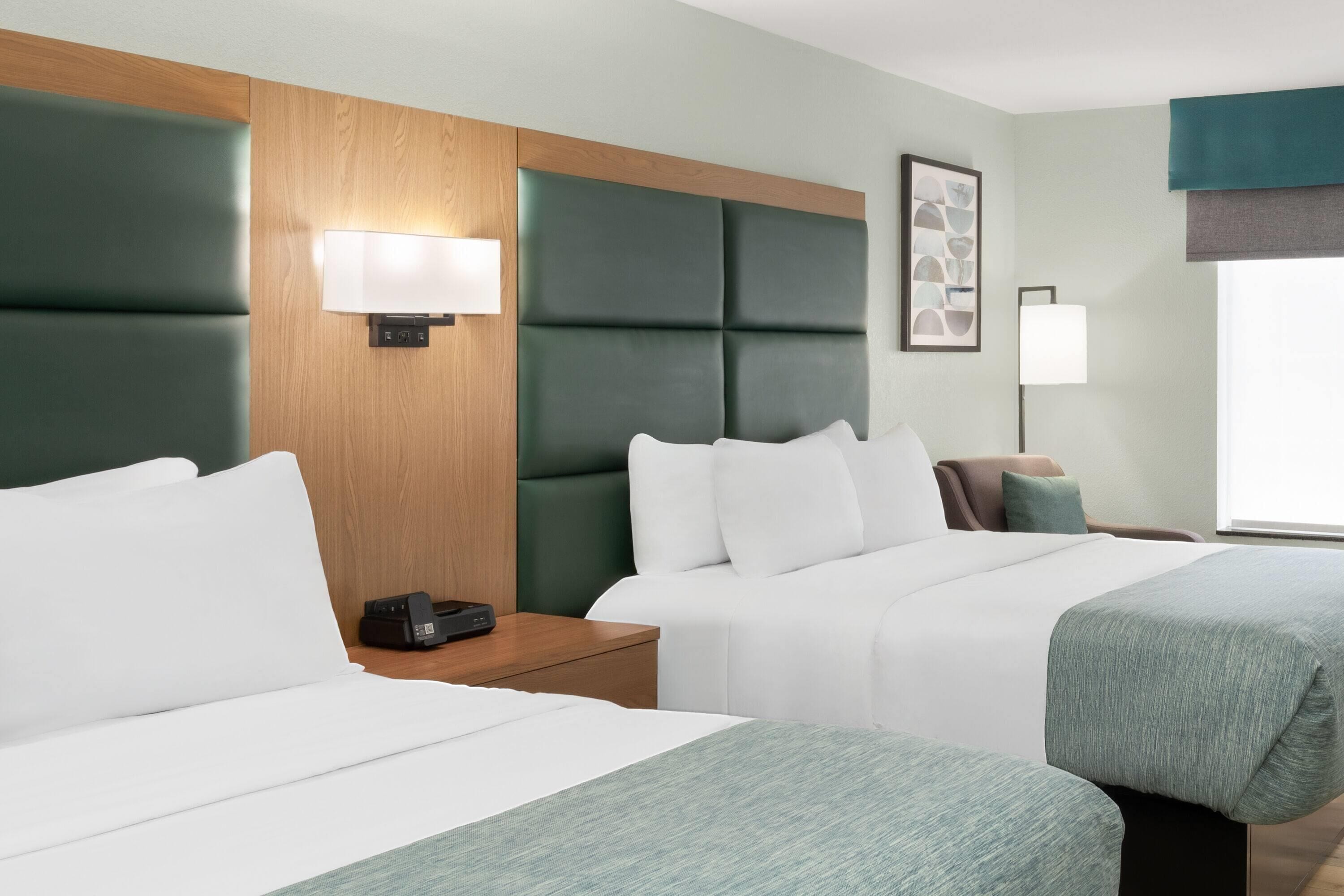 Habitación de hotel con dos camas, cabecero de madera, mesita de noche con lámpara y ventana con vistas.