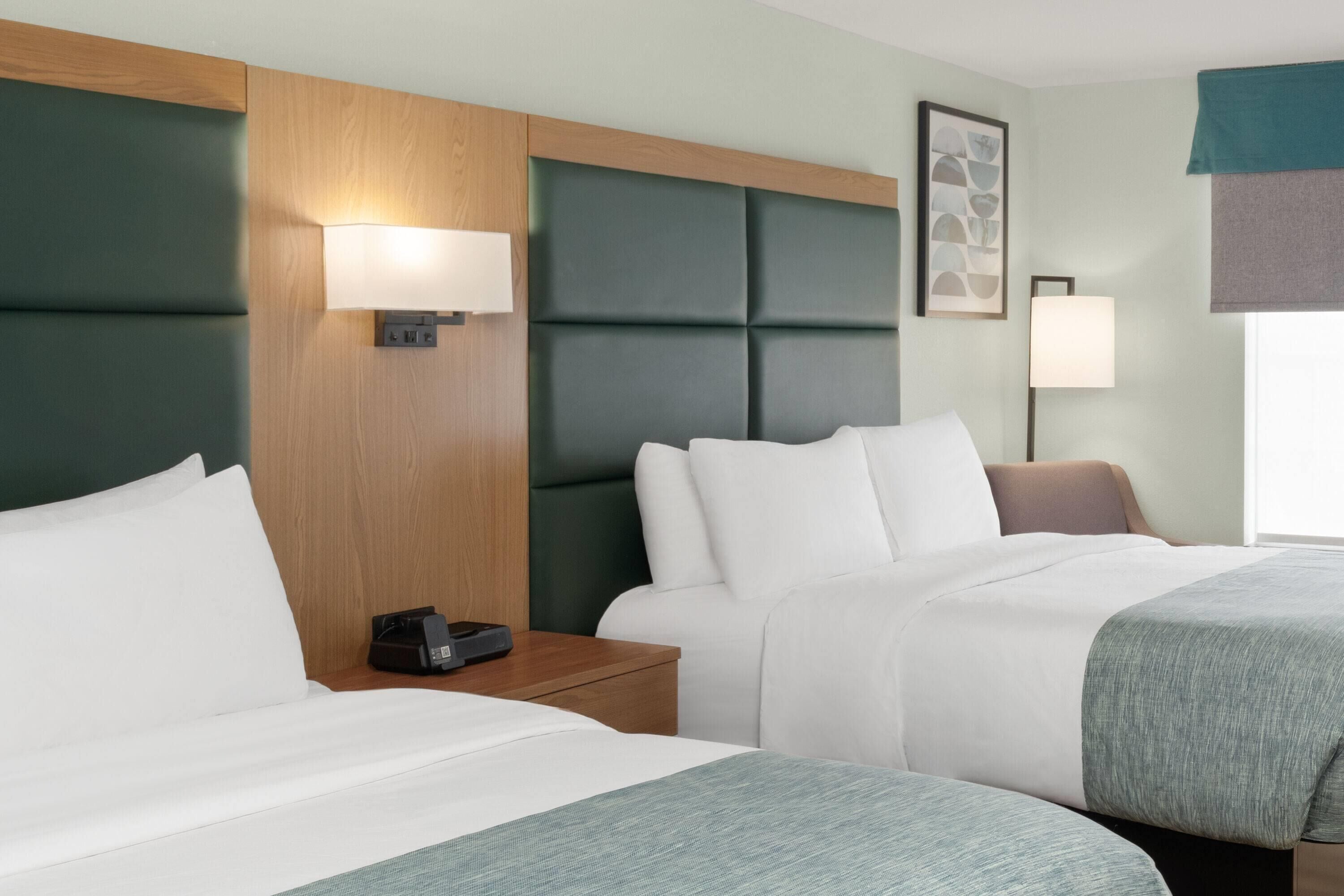A hotel room with two beds, a wooden headboard, a bedside table with a lamp, and a window with a view.