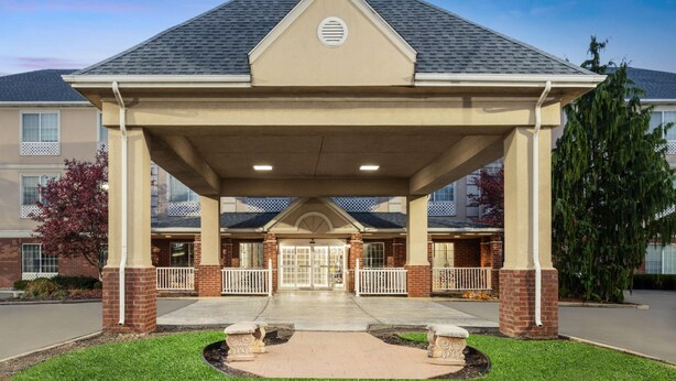 A covered entrance with a brick facade, a central door, and a small garden area with benches. - Wingate Suites By Wyndham Mansfield (Mansfield)
