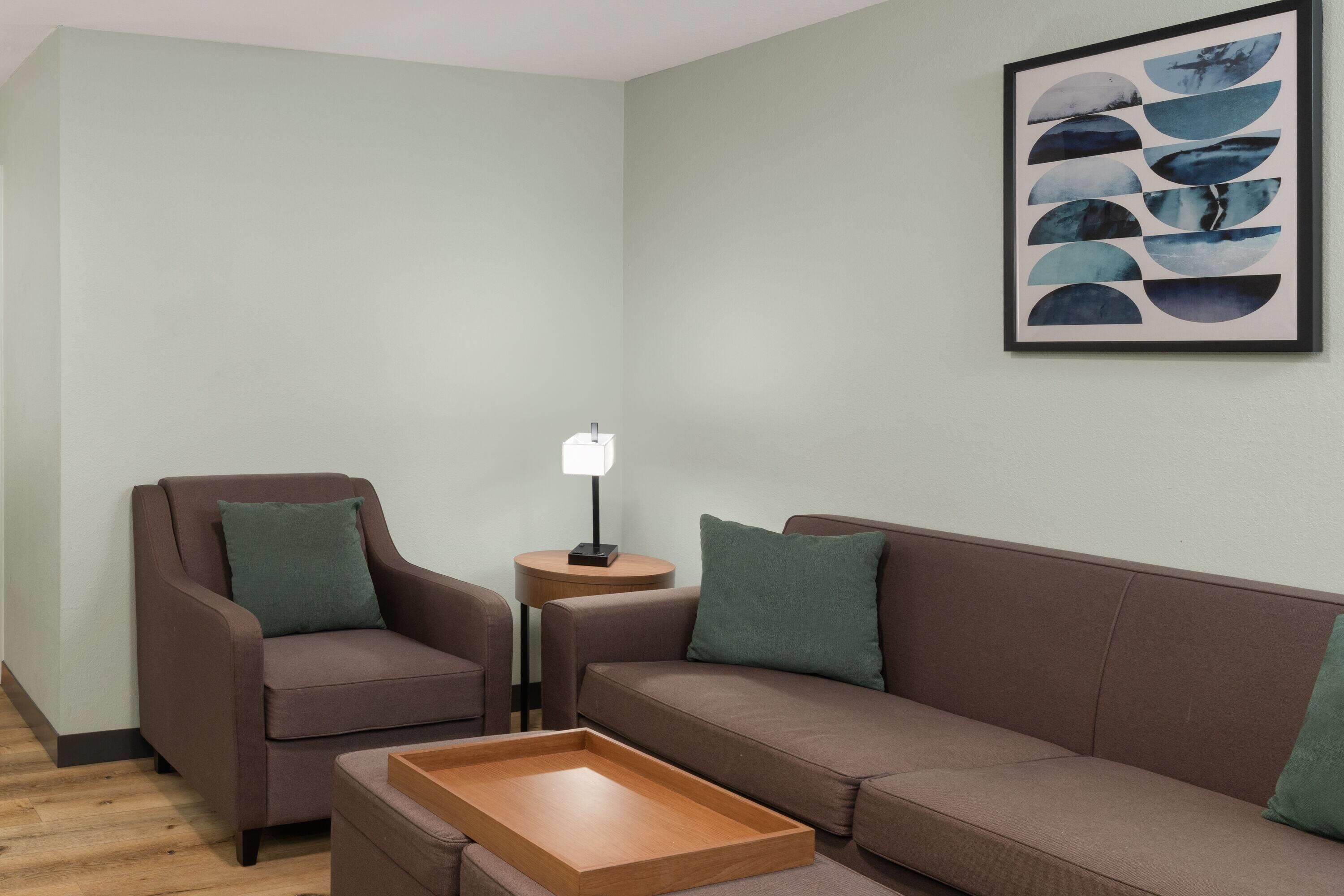 En moderne stue med en brun sofa, en lænestol i brunt, et træsidebord med en lampe, og et abstrakt kunstværk i ramme på væggen.