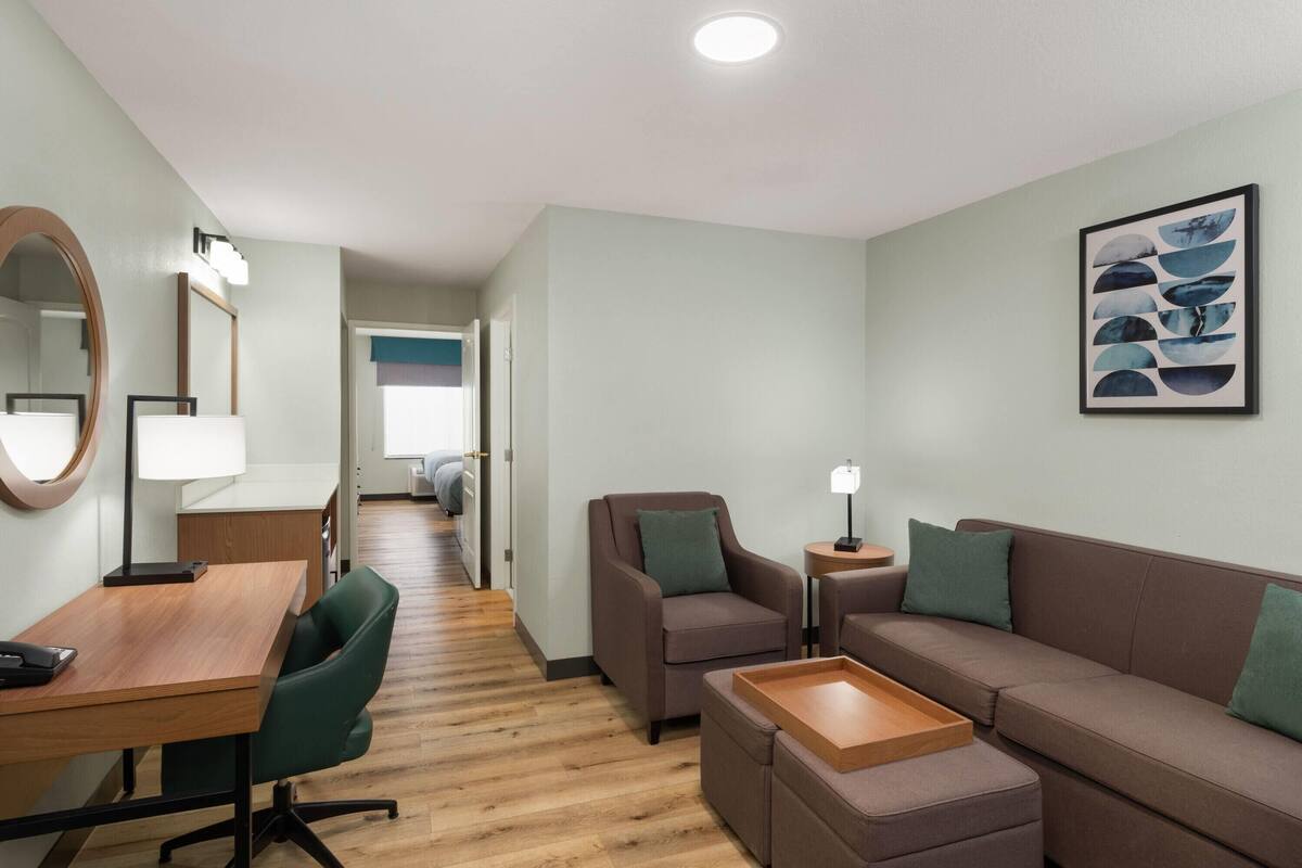Ein modernes Hotelzimmer mit einem Holztisch, einem grünen Bürostuhl, einem braunen Sofa und einem kleinen Nachttisch mit Lampe.
