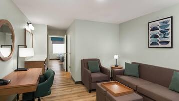 A modern hotel room with a wooden desk, green office chair, brown sofa, and a small side table with a lamp.