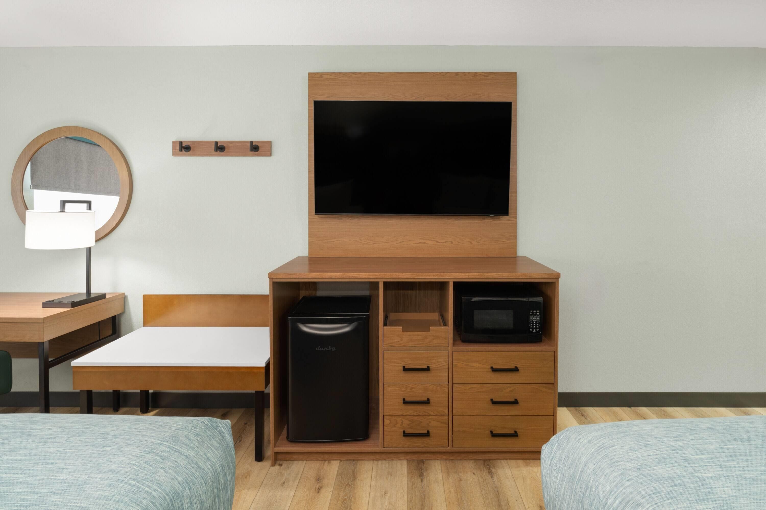 Quarto de hotel com cama, escrivaninha, espelho, televisão, micro-ondas e mini-geladeira.
