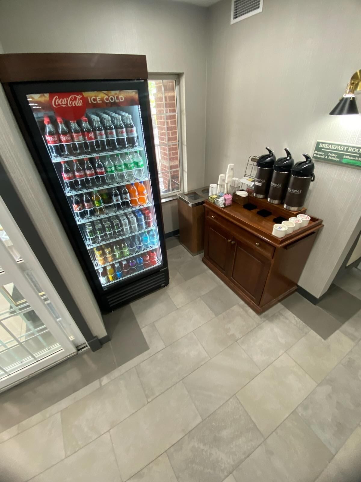 A Coca-Cola refrigerator stocked with various beverages, a coffee station with branded dispensers, and a sign indicating the breakfast room is open Monday to Friday, weekends, and holidays.