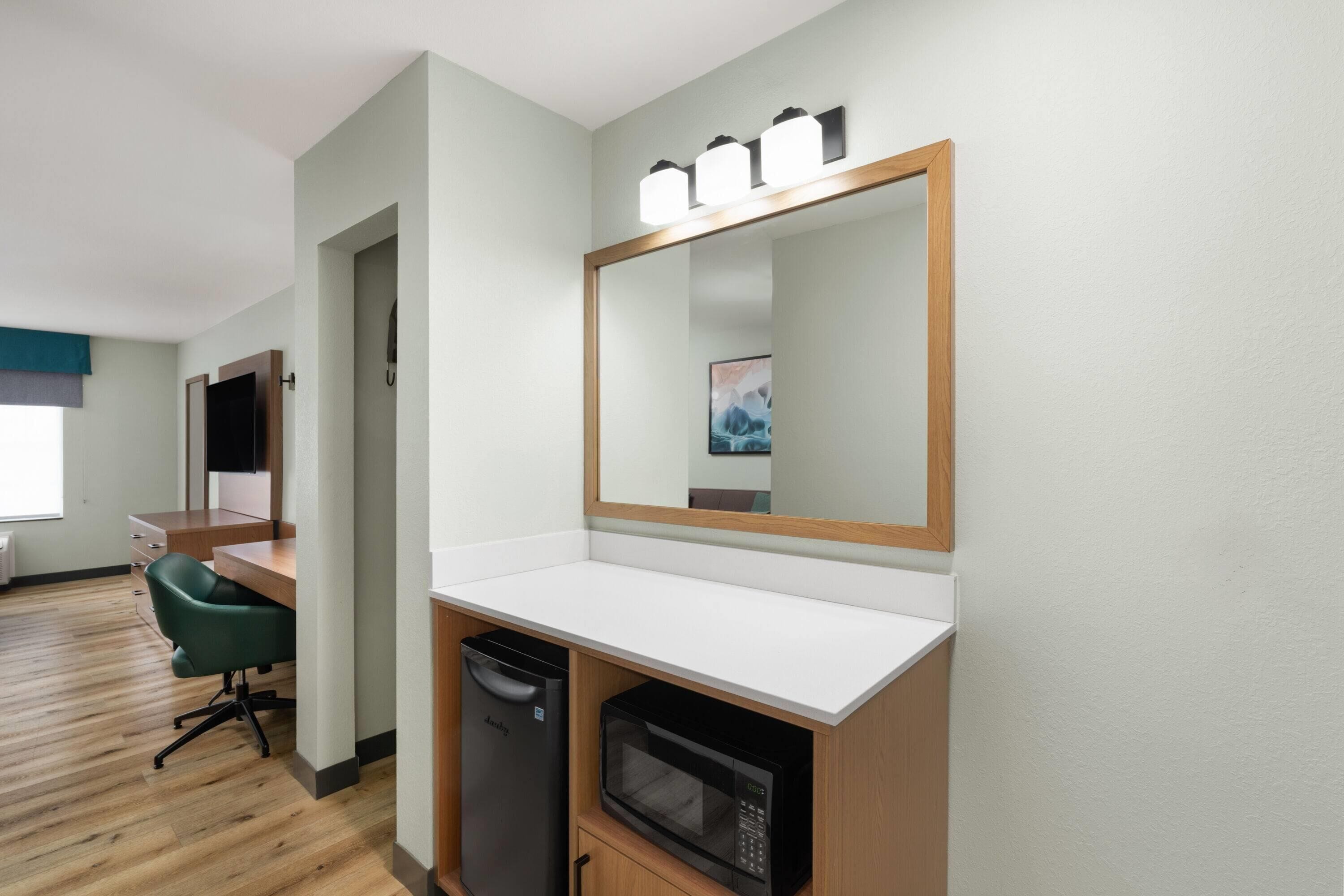 A hotel room with a wooden desk, a green office chair, a microwave, and a compact dishwasher.