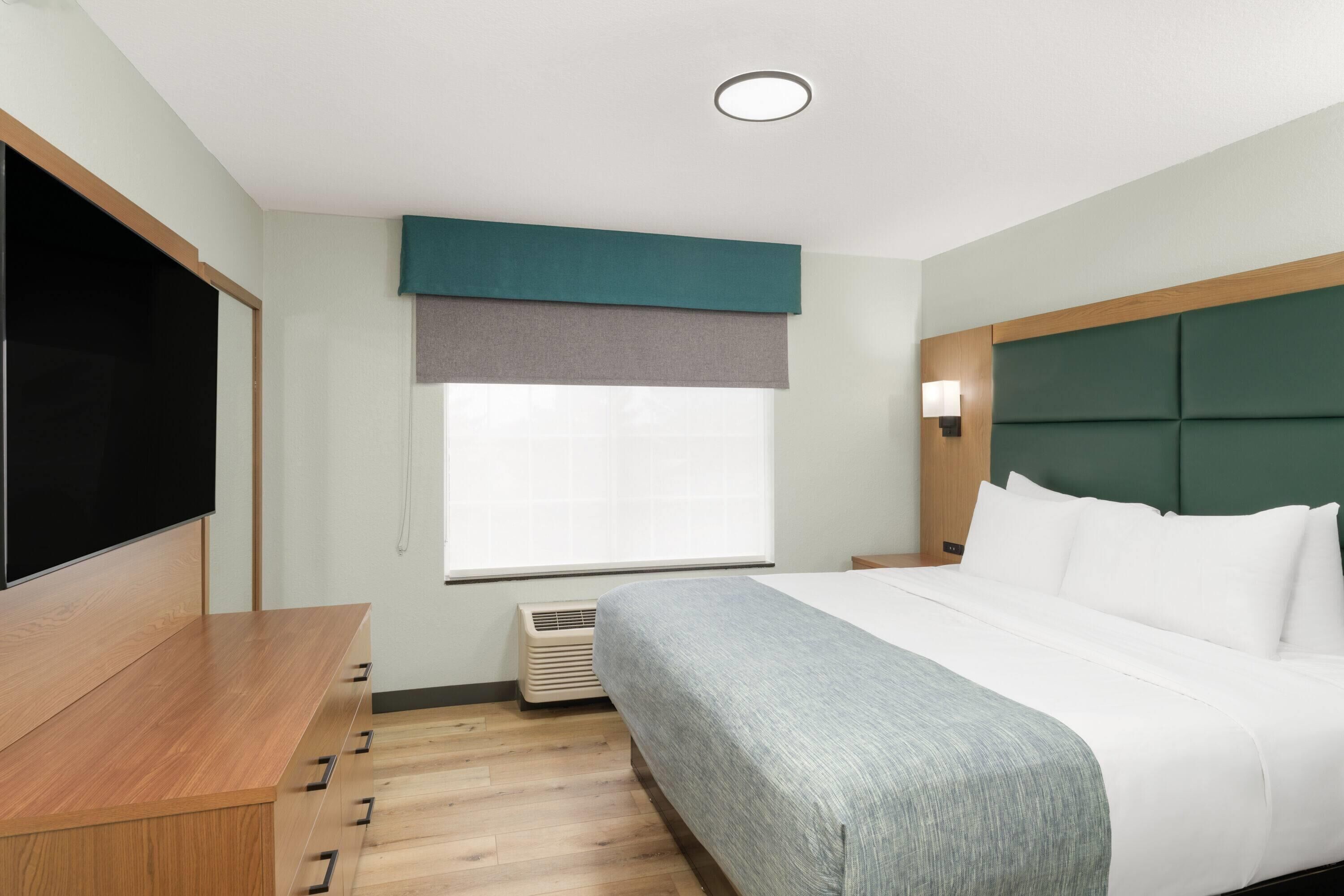 A hotel room with a bed, a flat-screen TV, a wooden desk, and a window with blinds.
