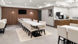 A modern dining area with a long table and chairs, a flat-screen TV mounted on the wall, and a coffee station with a microwave and coffee maker. - Wingate Suites By Wyndham Mansfield (Mansfield)