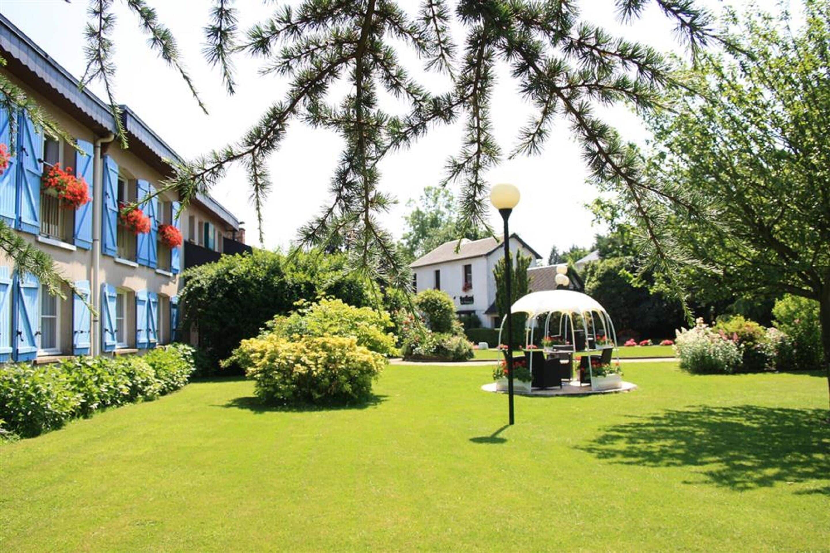 Un jardín cuidado con un pabellón, un edificio con contraventanas azules y una casa blanca al fondo.