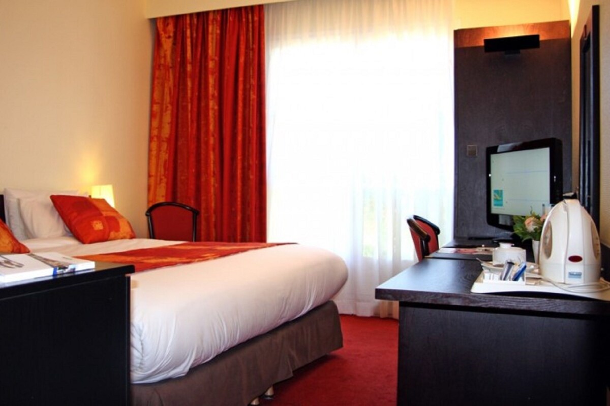 Ein Hotelzimmer mit Bett, Schreibtisch, Stühlen, Fernseher und einem Fenster mit Vorhängen.