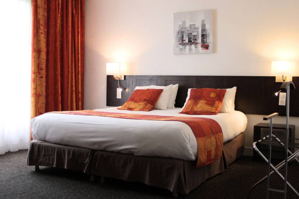 Ein Hotelzimmer mit einem großen Bett, zwei Nachttischlampen, einem Stadtbild an der Wand und einem Fenster mit Vorhängen.