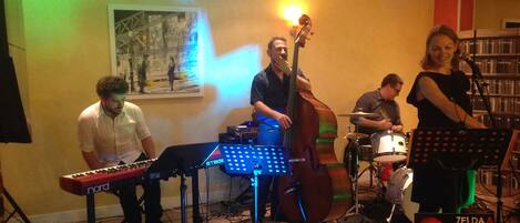 A jazz quartet performing in a room with a tiled floor, a painting on the wall, and a bookshelf in the background.