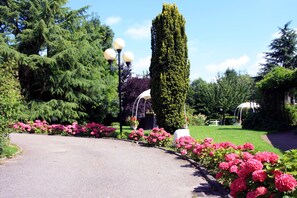 A garden pathway with a central circular flower bed, surrounded by tall trees and a tall, narrow tree. - La Bertelière, The Originals Relais Rouen (Saint-Martin-du-Vivier)