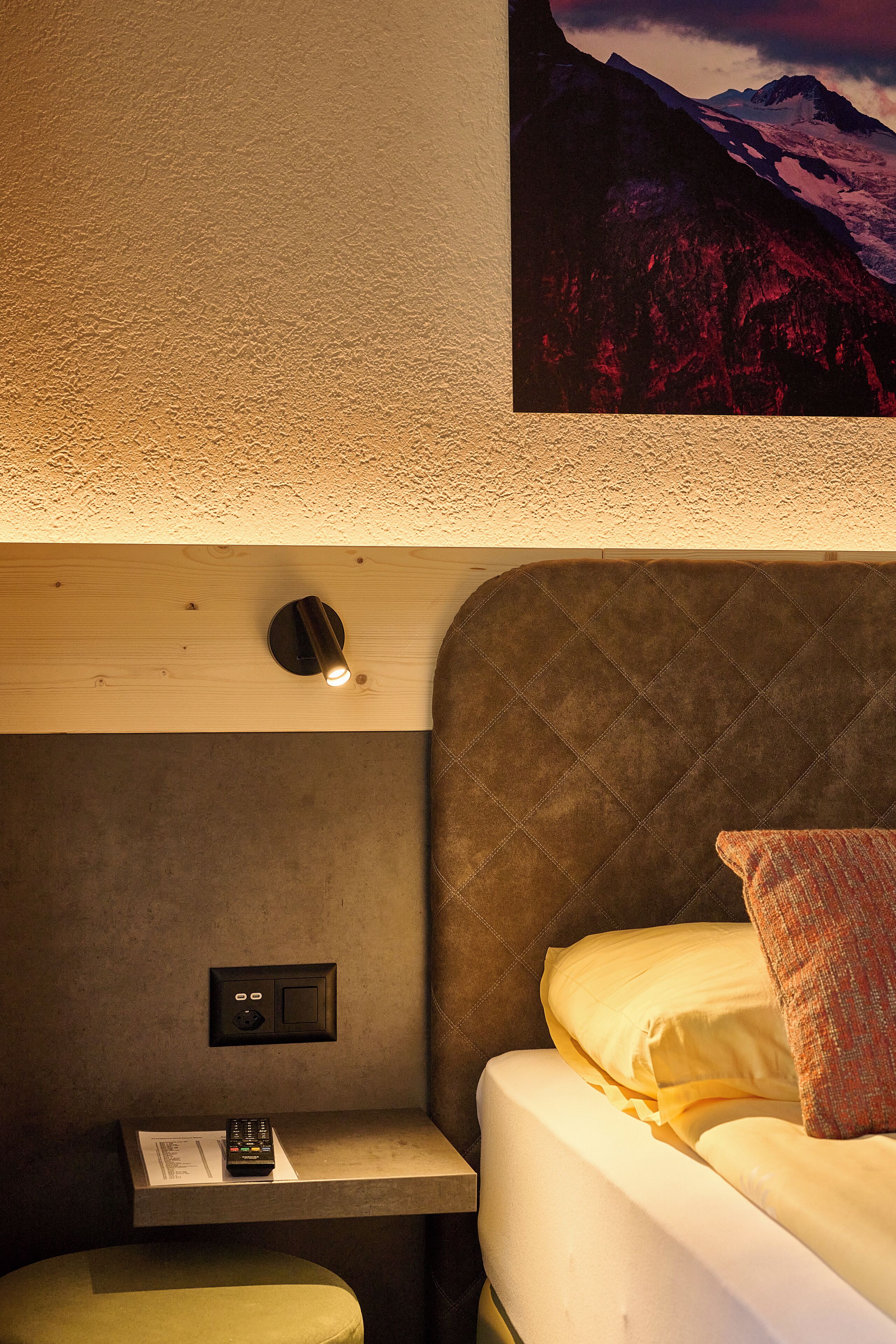 Habitación de hotel con cama, mesita de noche con control remoto, estante pequeño y lámpara de pared.