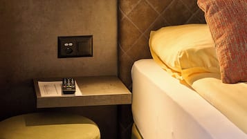 Habitación de hotel con cama, mesita de noche con control remoto, estante pequeño y lámpara de pared.