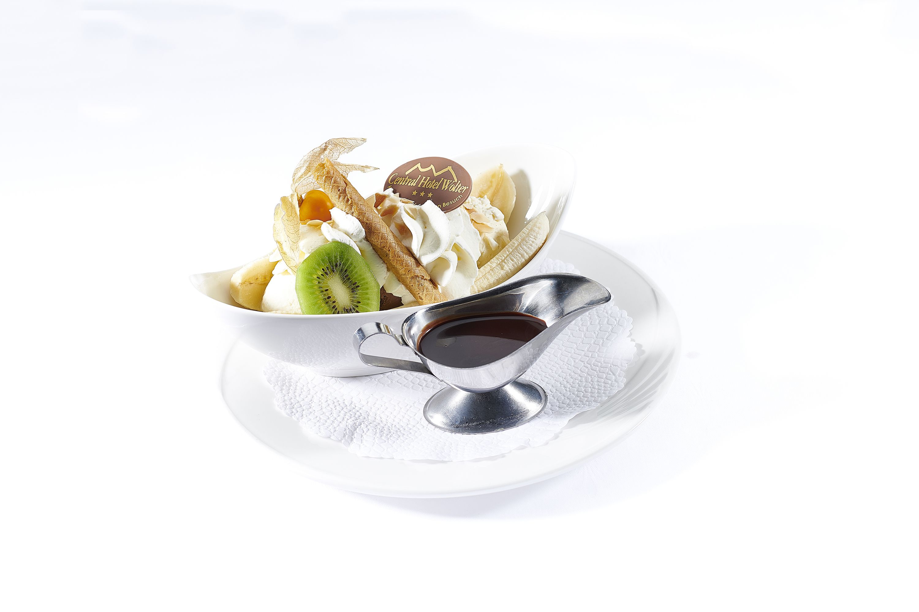 Un postre con crema batida, rodajas de plátano y un trozo de chocolate sobre un plato blanco, junto con una cuchara y una taza pequeña de salsa.
