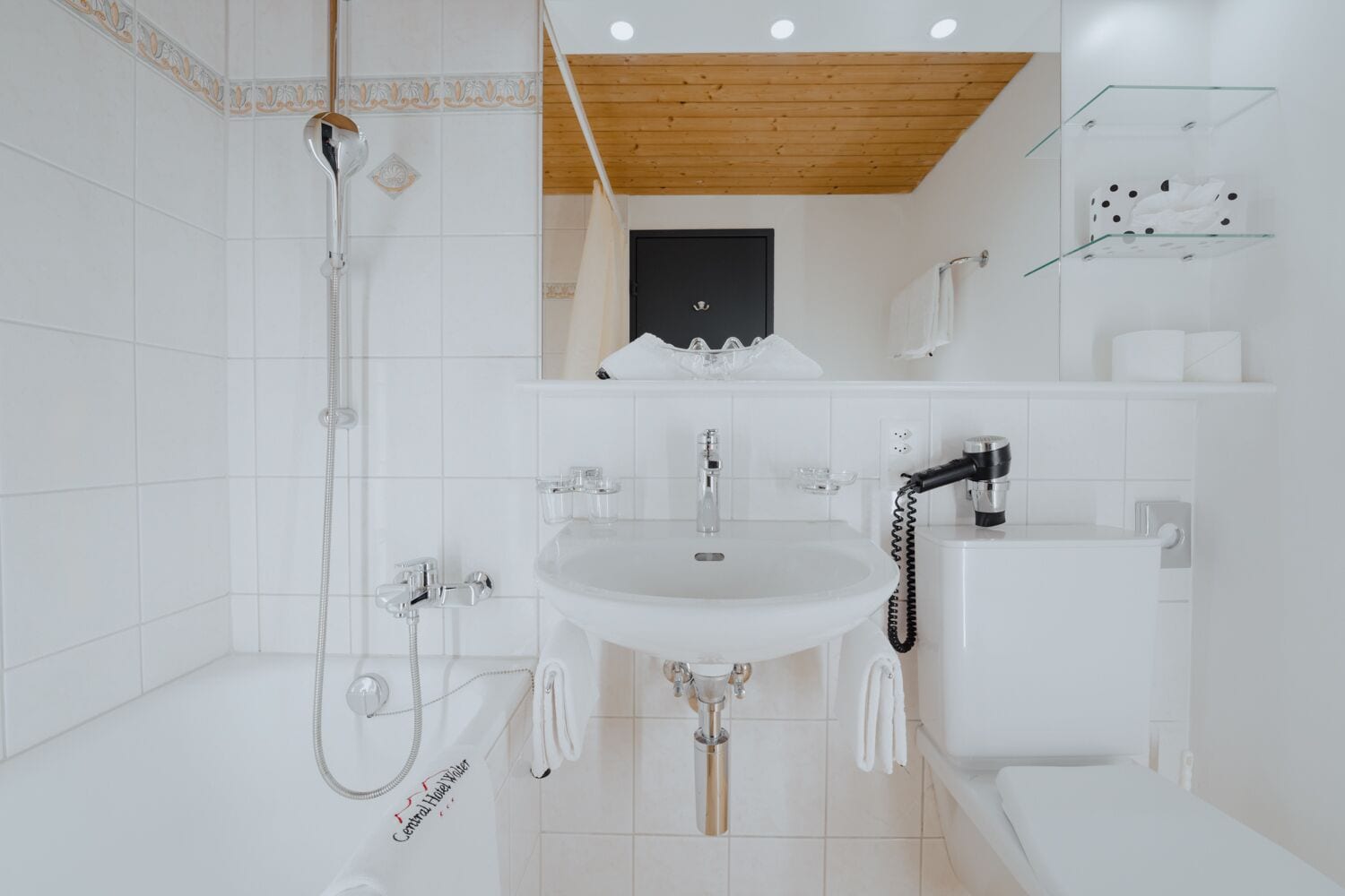 A modern bathroom with a white sink, toilet, and shower. There is a towel rack and a glass shelf with toiletries.