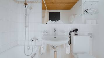 Un baño moderno con lavamanos, inodoro y ducha blancos. Hay un perchero y un estante de vidrio con artículos de aseo.