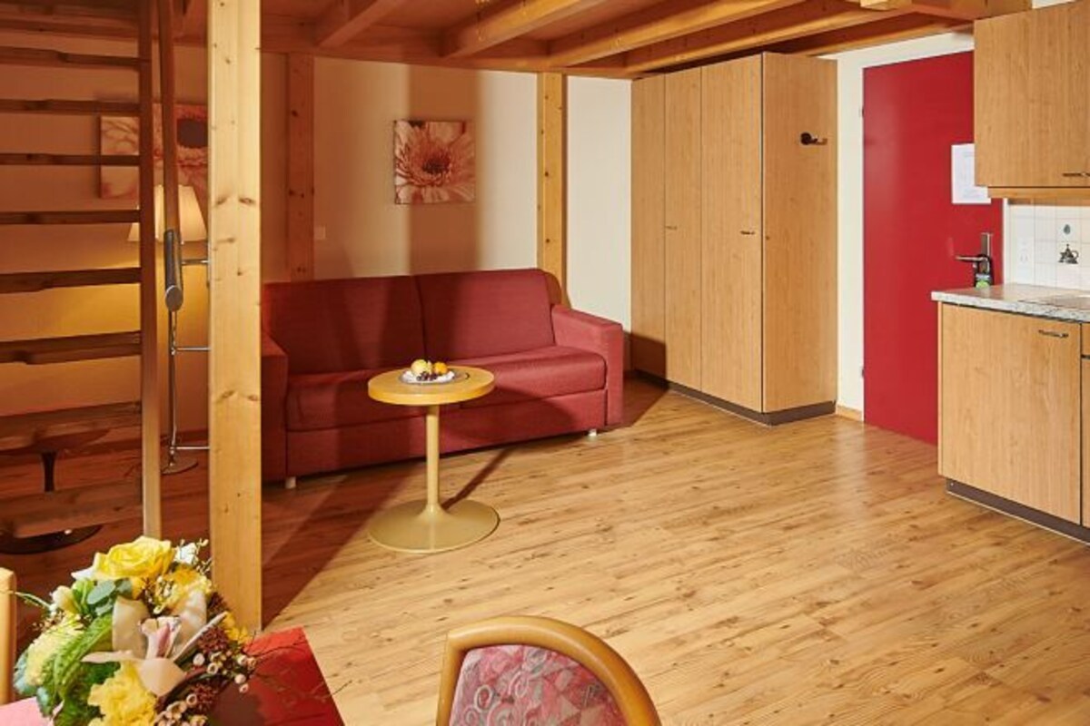 A cozy room with a wooden floor, a red sofa, a small round table, and a kitchenette with wooden cabinets.