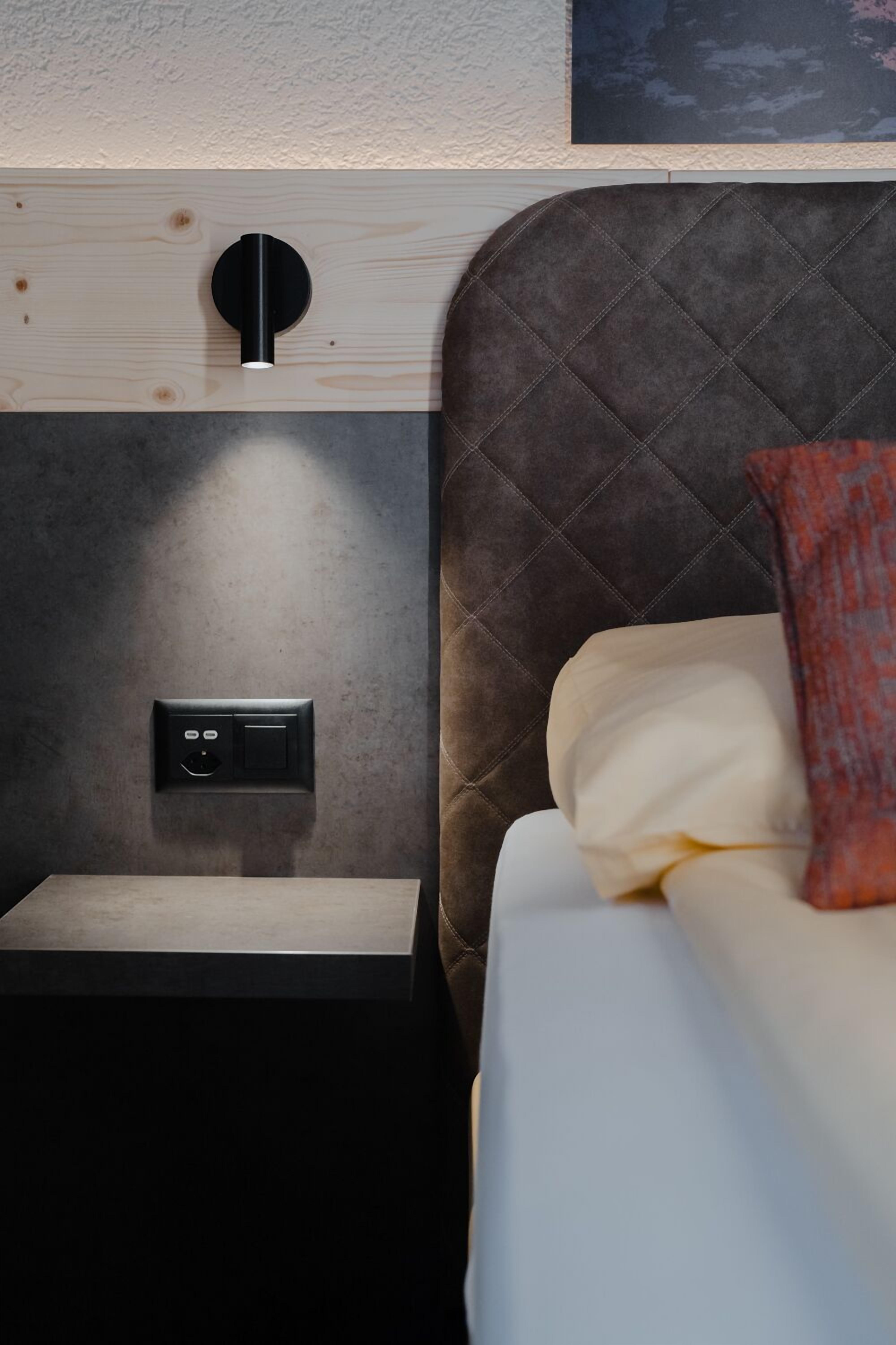 A modern hotel room with a bed, a nightstand, a wall-mounted control panel, and a quilted headboard.