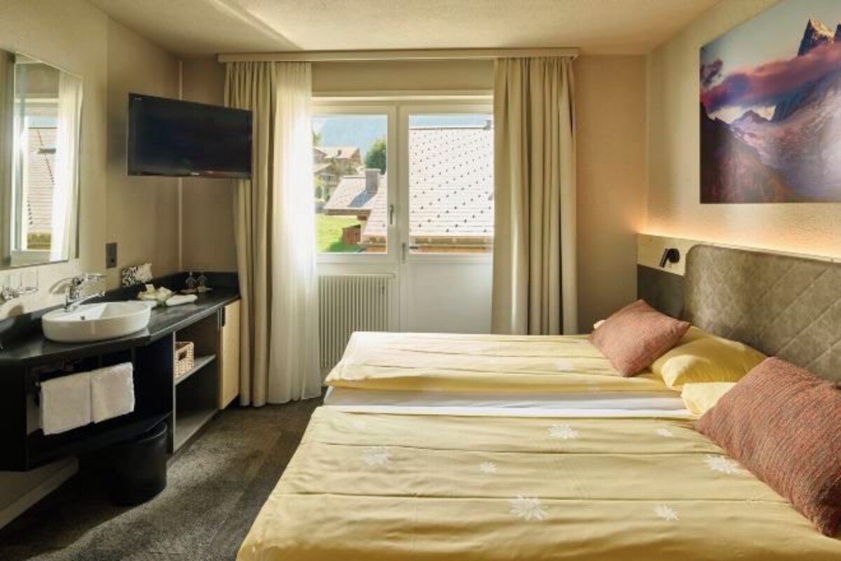 A hotel room with a bed, a TV mounted on the wall, a bathroom with a sink and mirror, and a window with curtains.