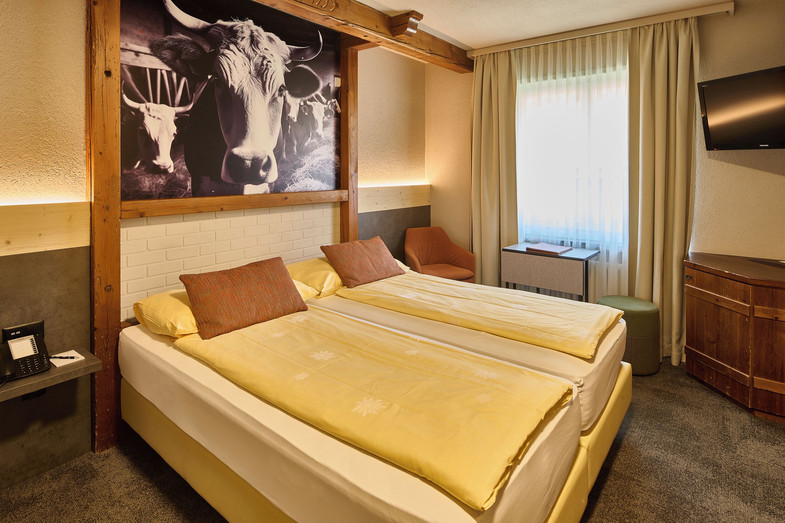 Habitación de hotel con dos camas, cabecera de madera, televisor de pantalla plana, una mesita y un cuadro de un toro.