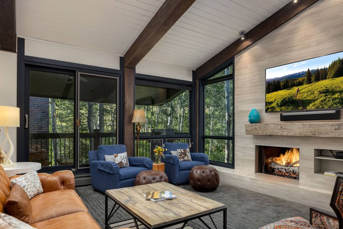 A modern living room with leather sofas, a wooden coffee table, and a fireplace. Large windows offer a view of a forested area.