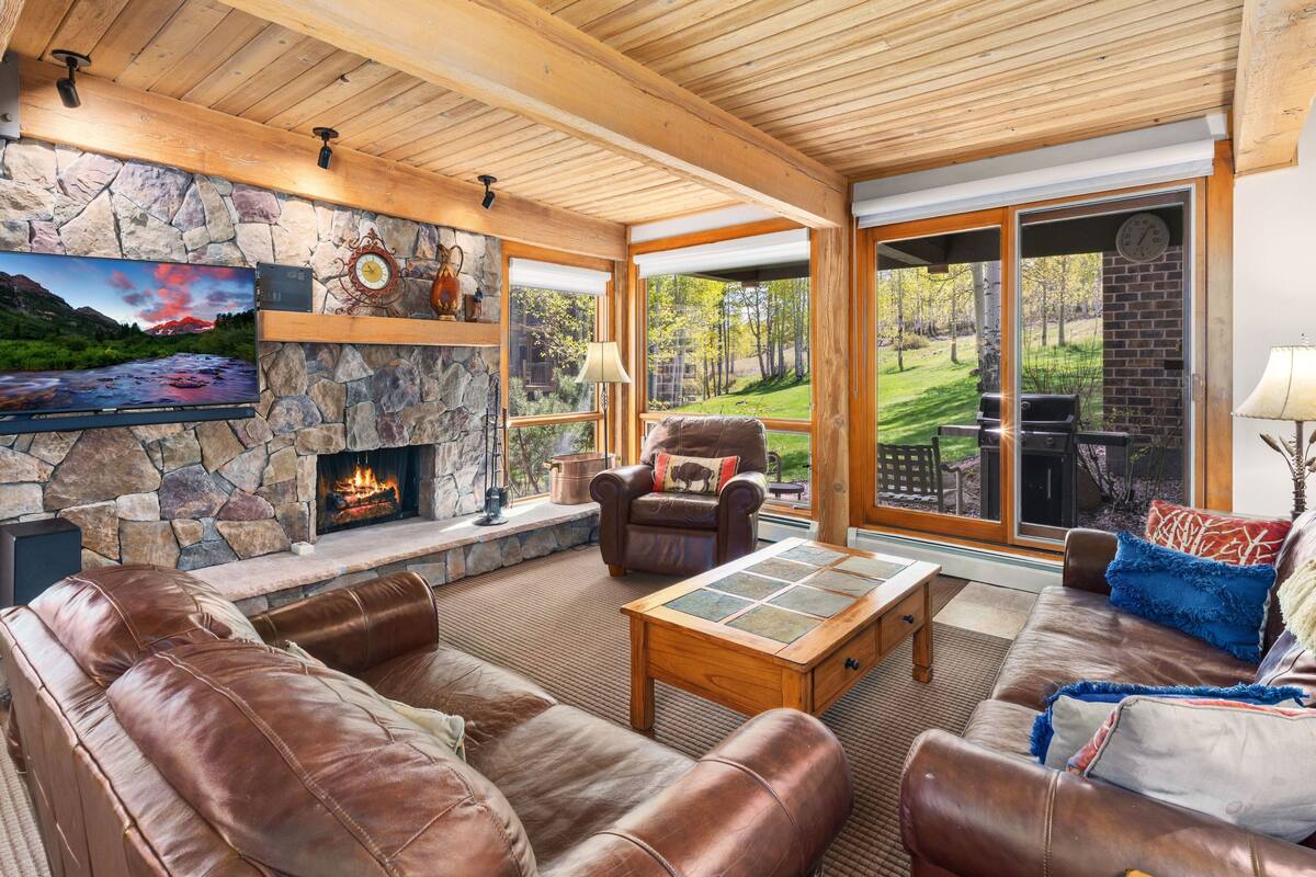 A cozy living room with a stone fireplace, leather sofas, and a large window offering a view of the outdoors.