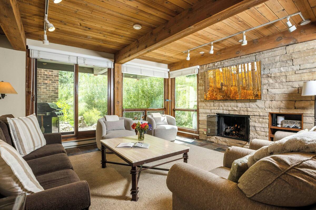 A spacious living room with a stone fireplace, large windows, and a wooden ceiling.
