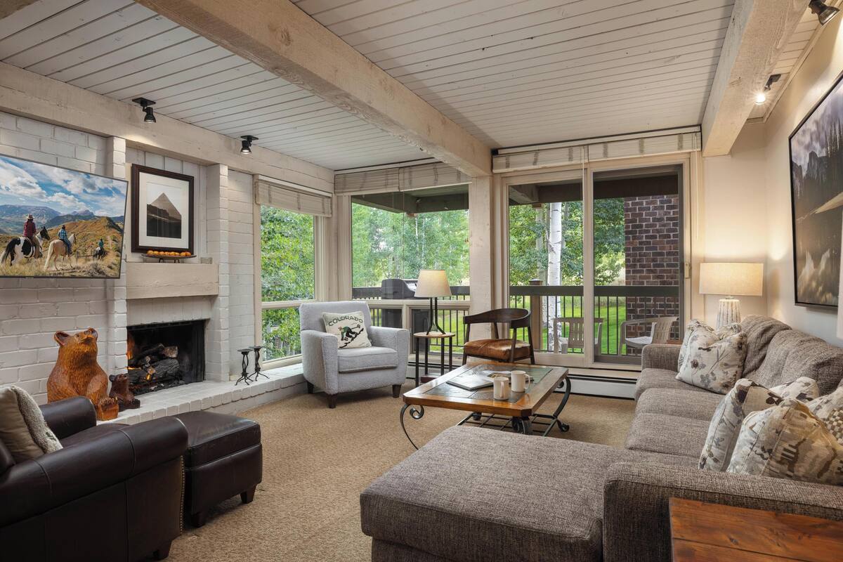 A cozy living room with a fireplace, comfortable seating, and large windows offering a view of the outdoors.
