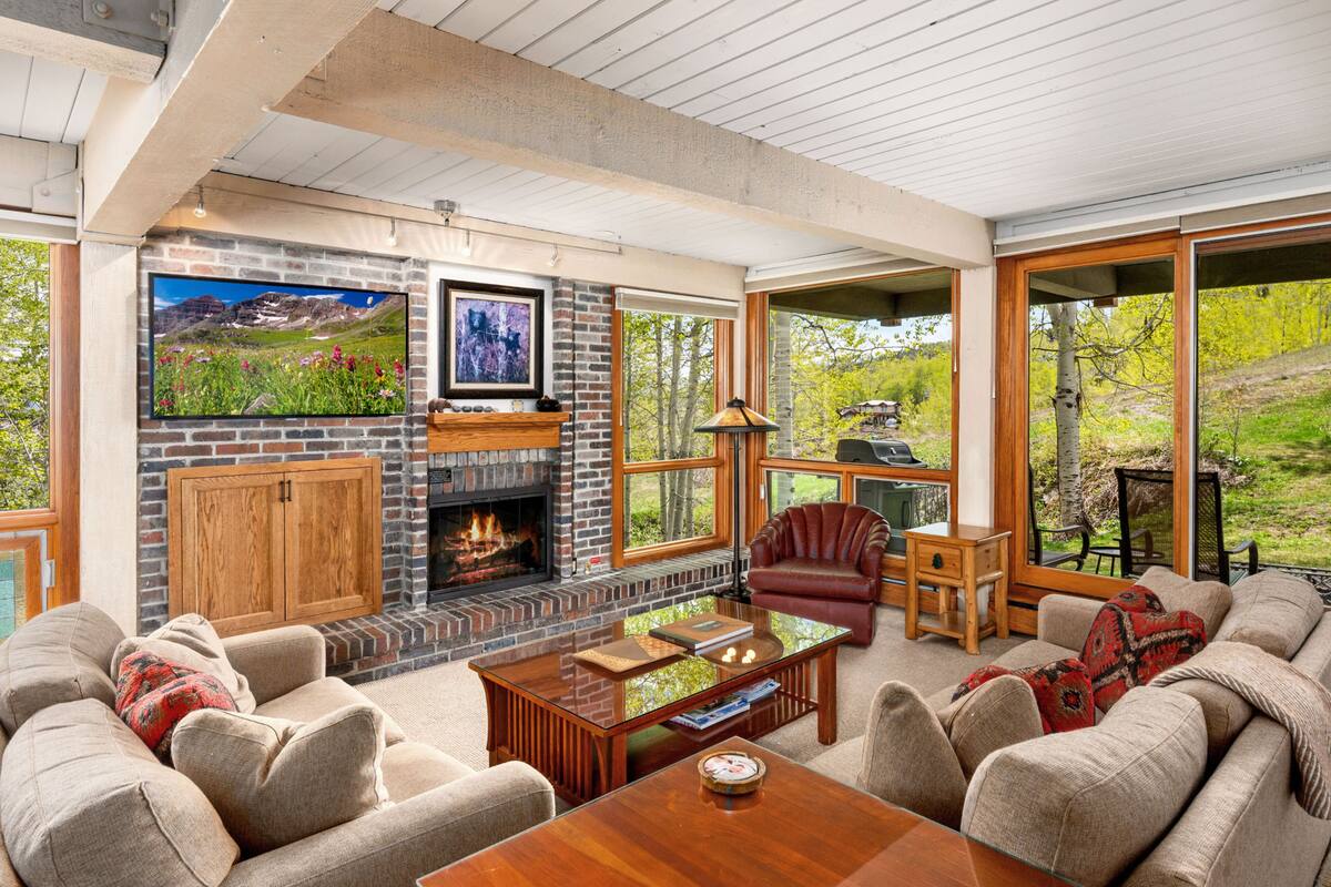 A cozy living room with a fireplace, comfortable seating, and large windows offering a view of the outdoors.