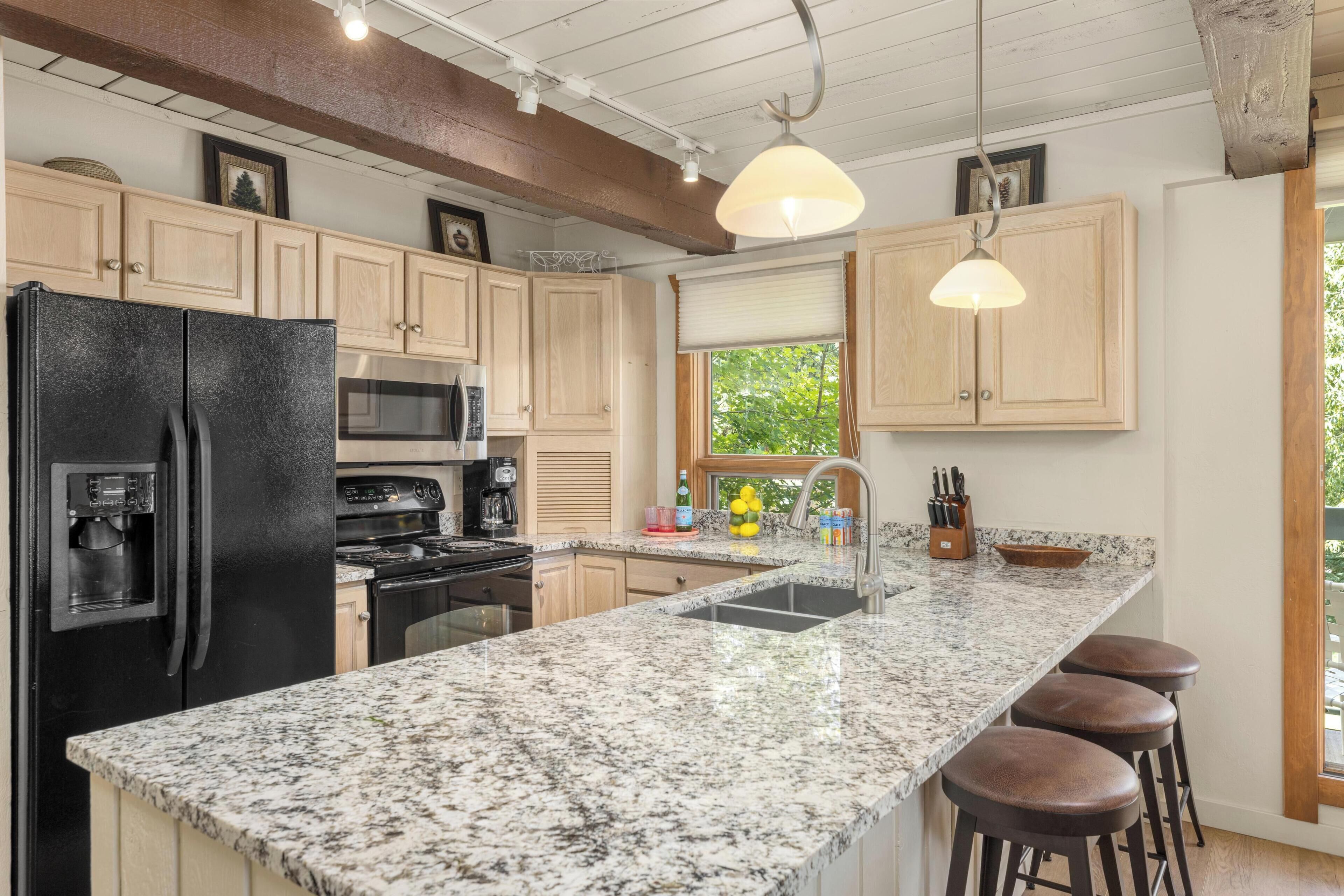 A kitchen with a granite countertop, stainless steel appliances, wooden cabinets, a window with a view, and bar stools.
