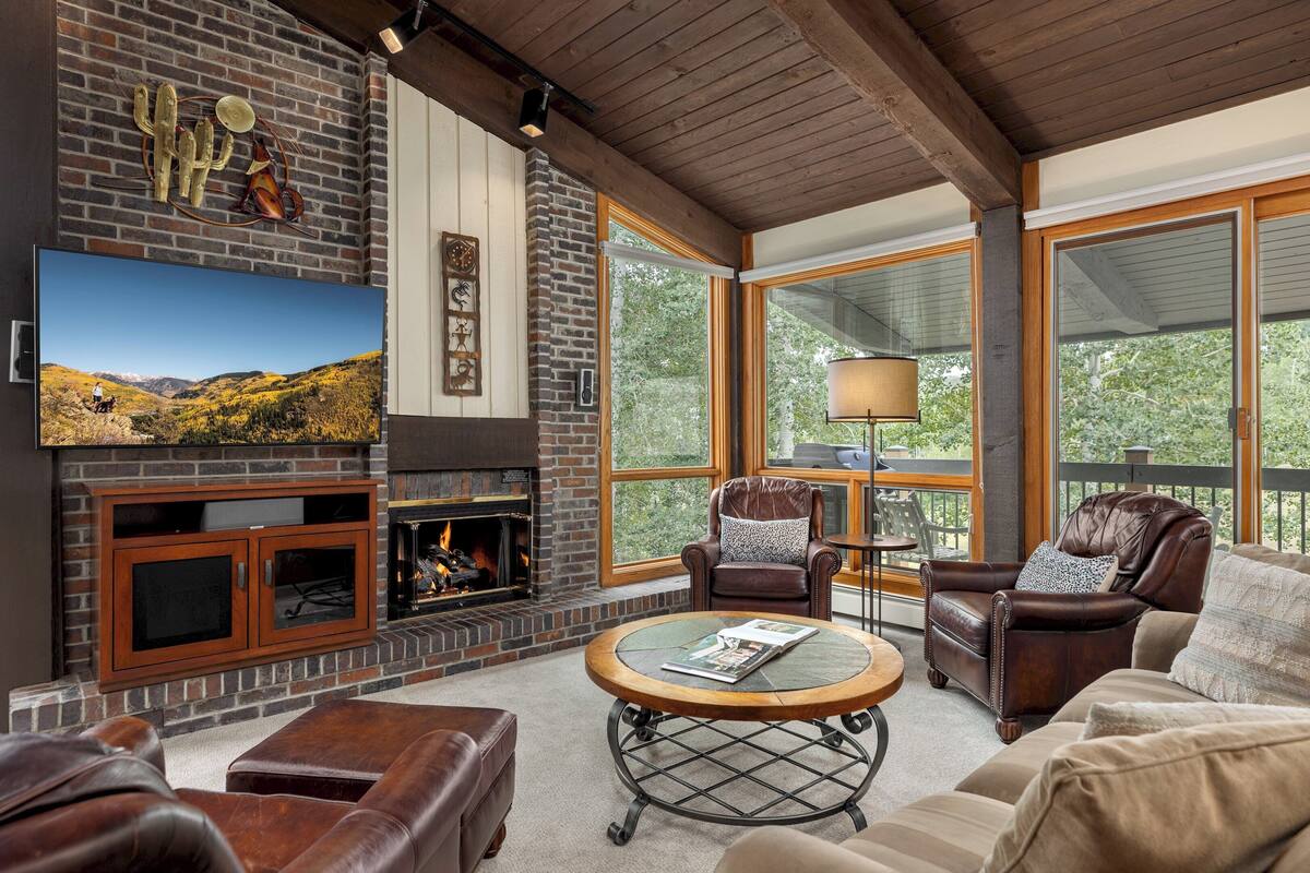 A cozy living room with a fireplace, leather furniture, and large windows offering a view of the outdoors.