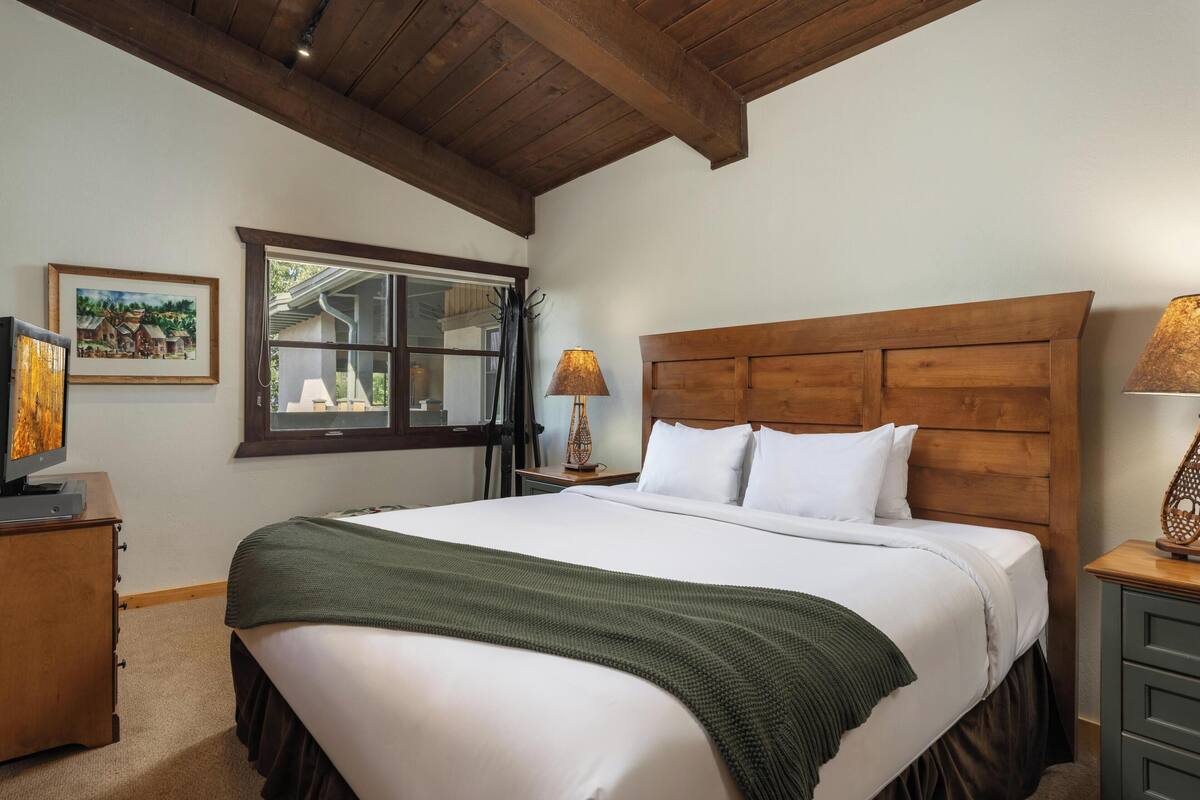 A neatly made bed with a wooden headboard, a green throw blanket, and white bedding. There is a wooden nightstand with a lamp and a framed picture on the wall.