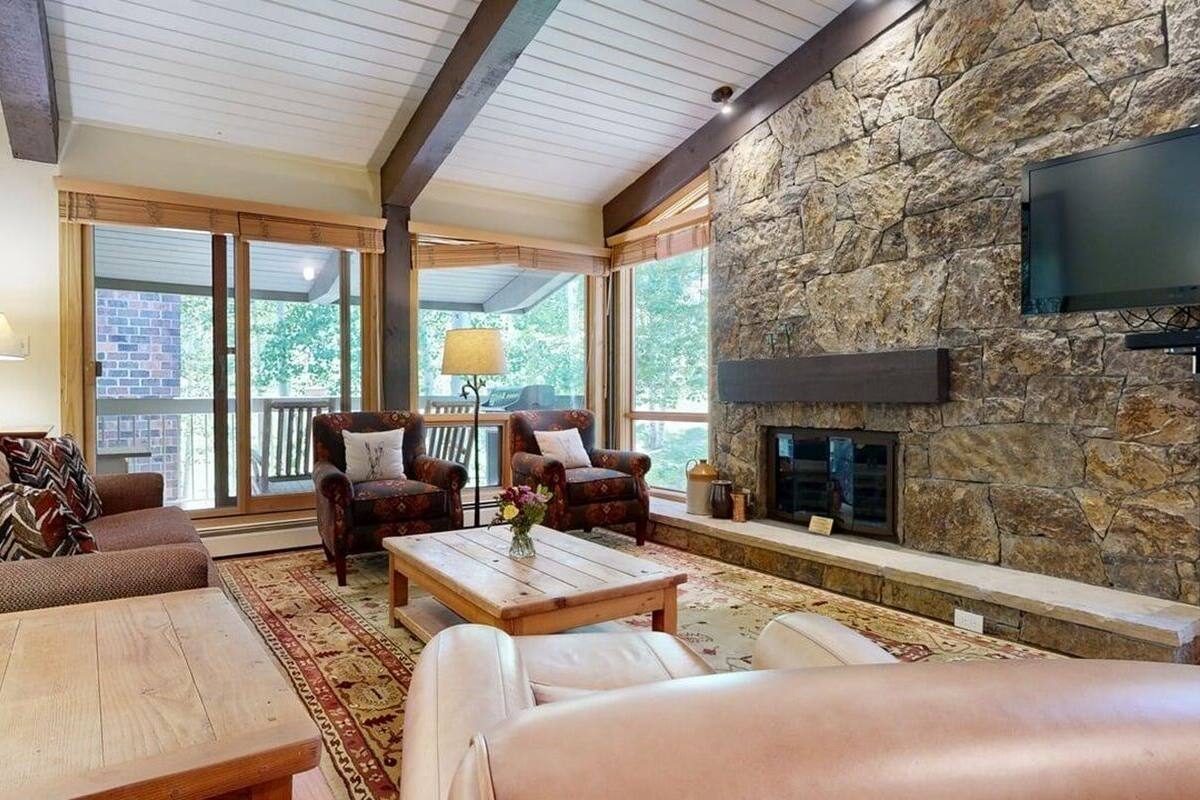 A spacious living room with a stone fireplace, comfortable seating, and large windows offering a view of greenery.