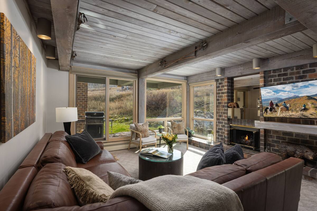 A cozy living room with a leather sofa, a fireplace, and large windows offering a view of the outdoors.