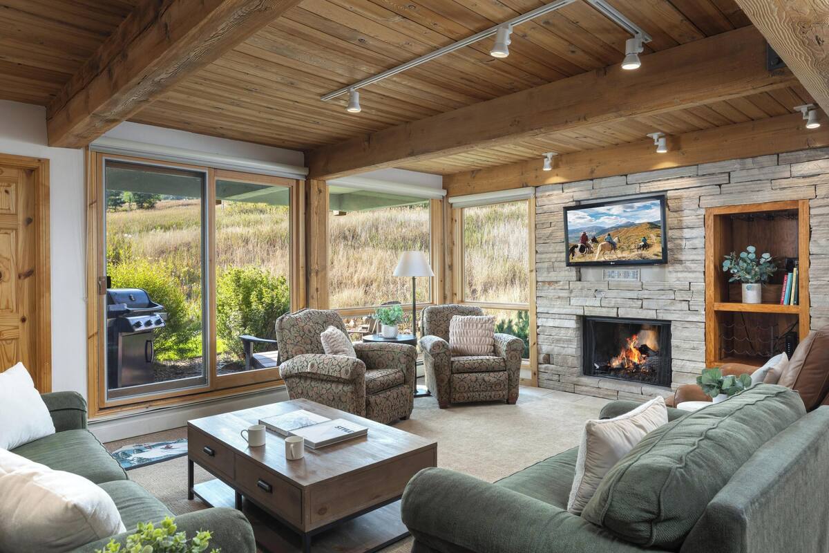 A cozy living room with a fireplace, comfortable seating, and large windows offering a view of the outdoors.