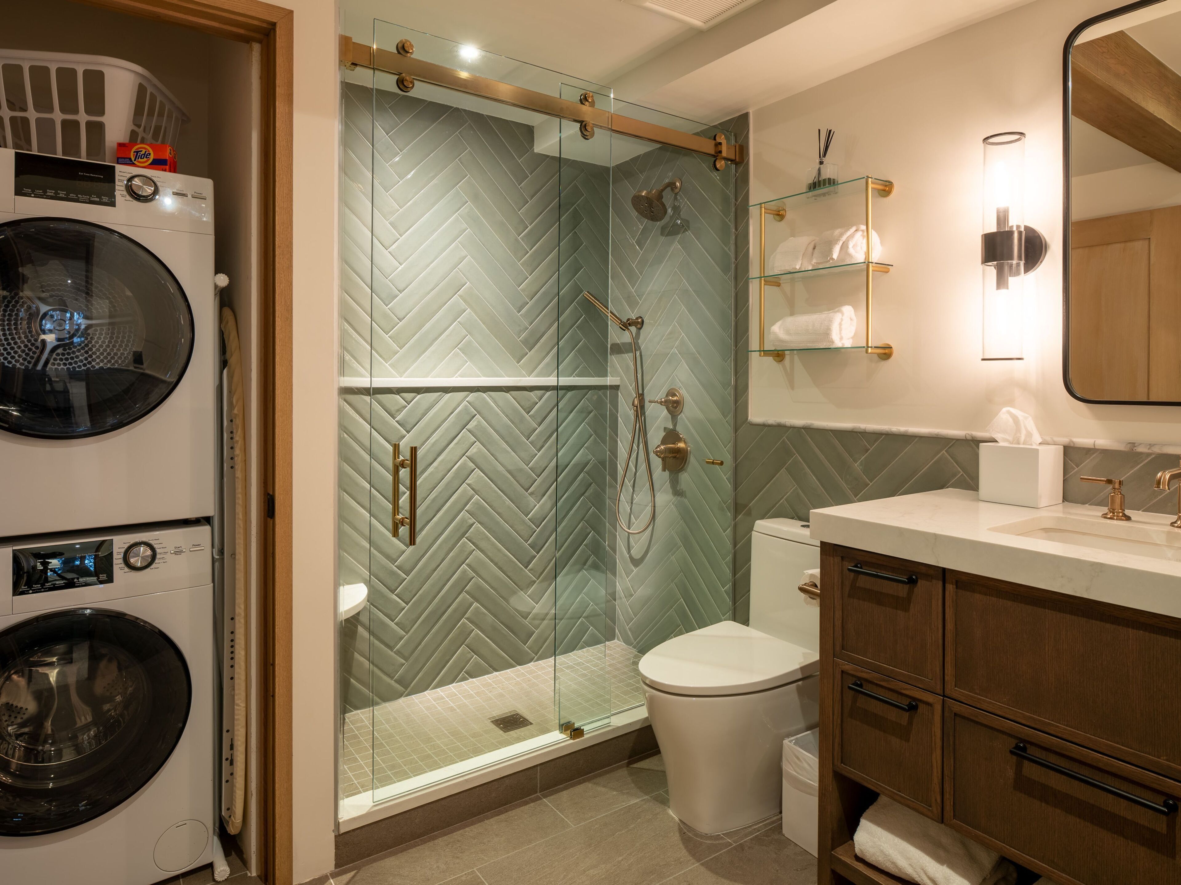 A modern bathroom with a glass shower enclosure, a built-in washing machine, and a wooden vanity with a mirror.