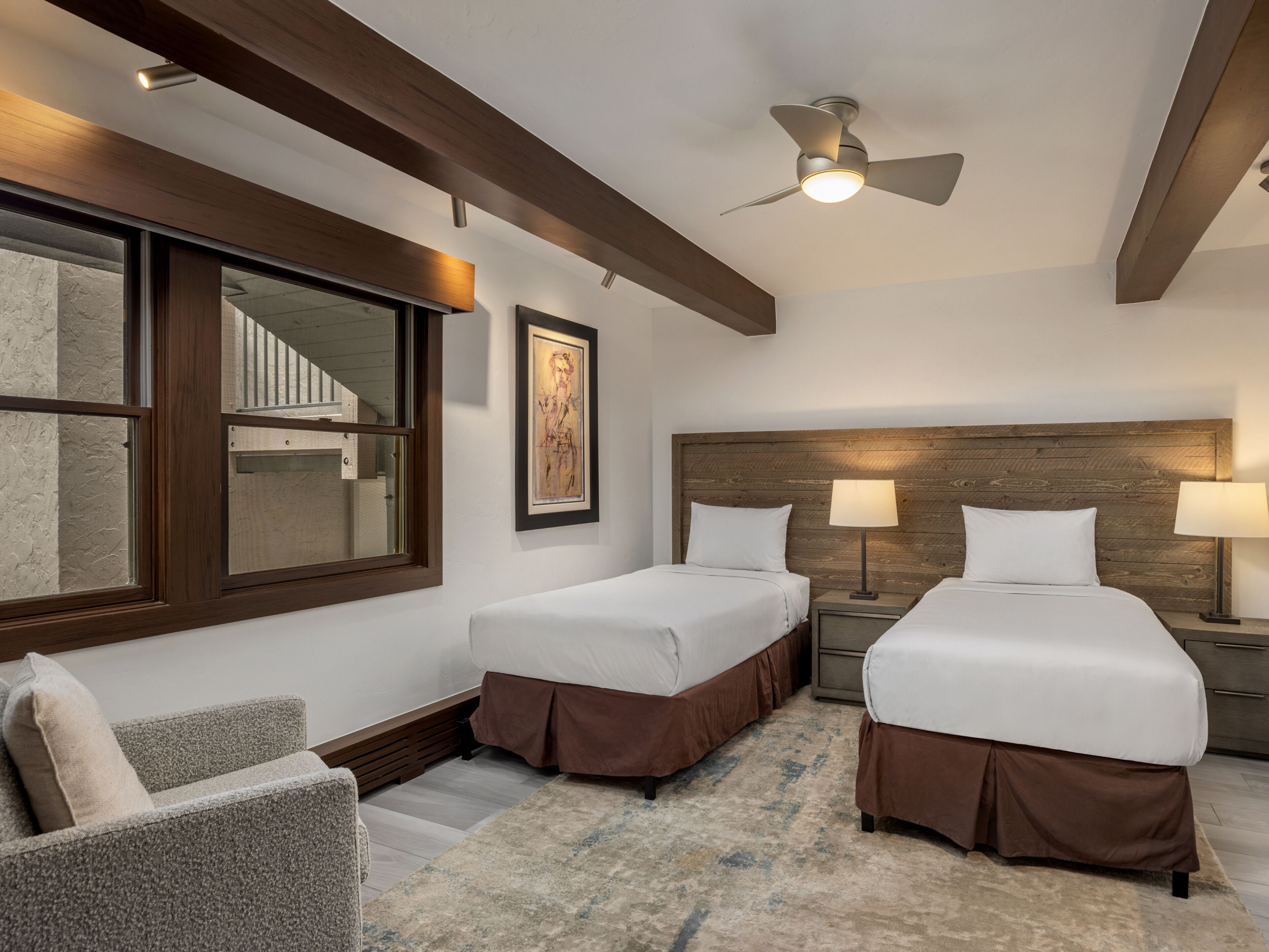 A hotel room with two beds, a chair, a framed picture, a ceiling fan, and a window with blinds.