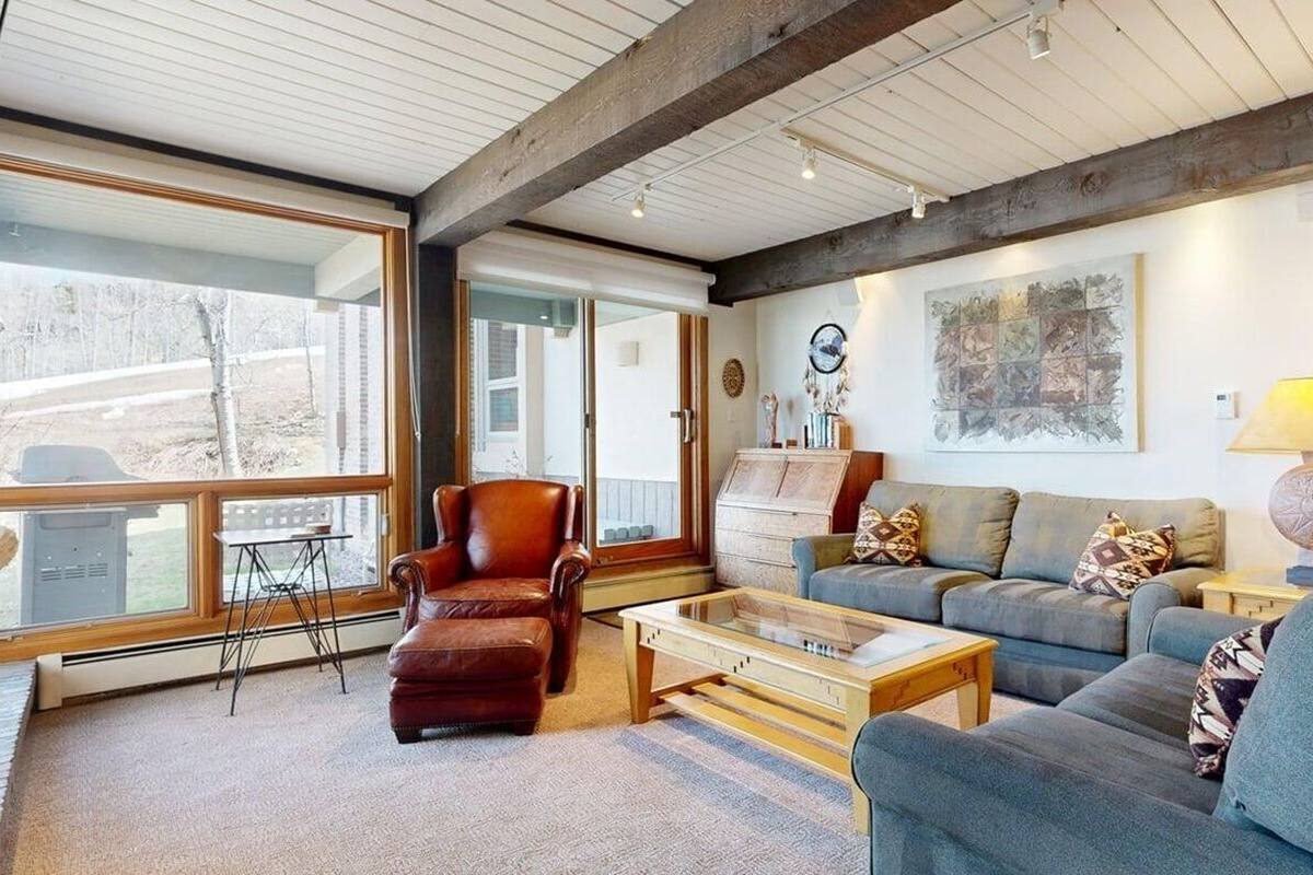 A living room with a sofa, armchair, coffee table, and a lamp. There is a window with a view of trees and a bench outside.