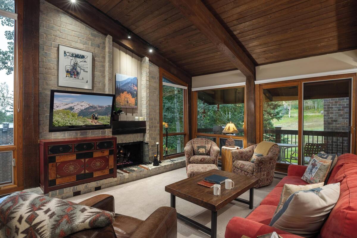 A cozy living room with a fireplace, comfortable seating, and large windows offering a view of the outdoors.