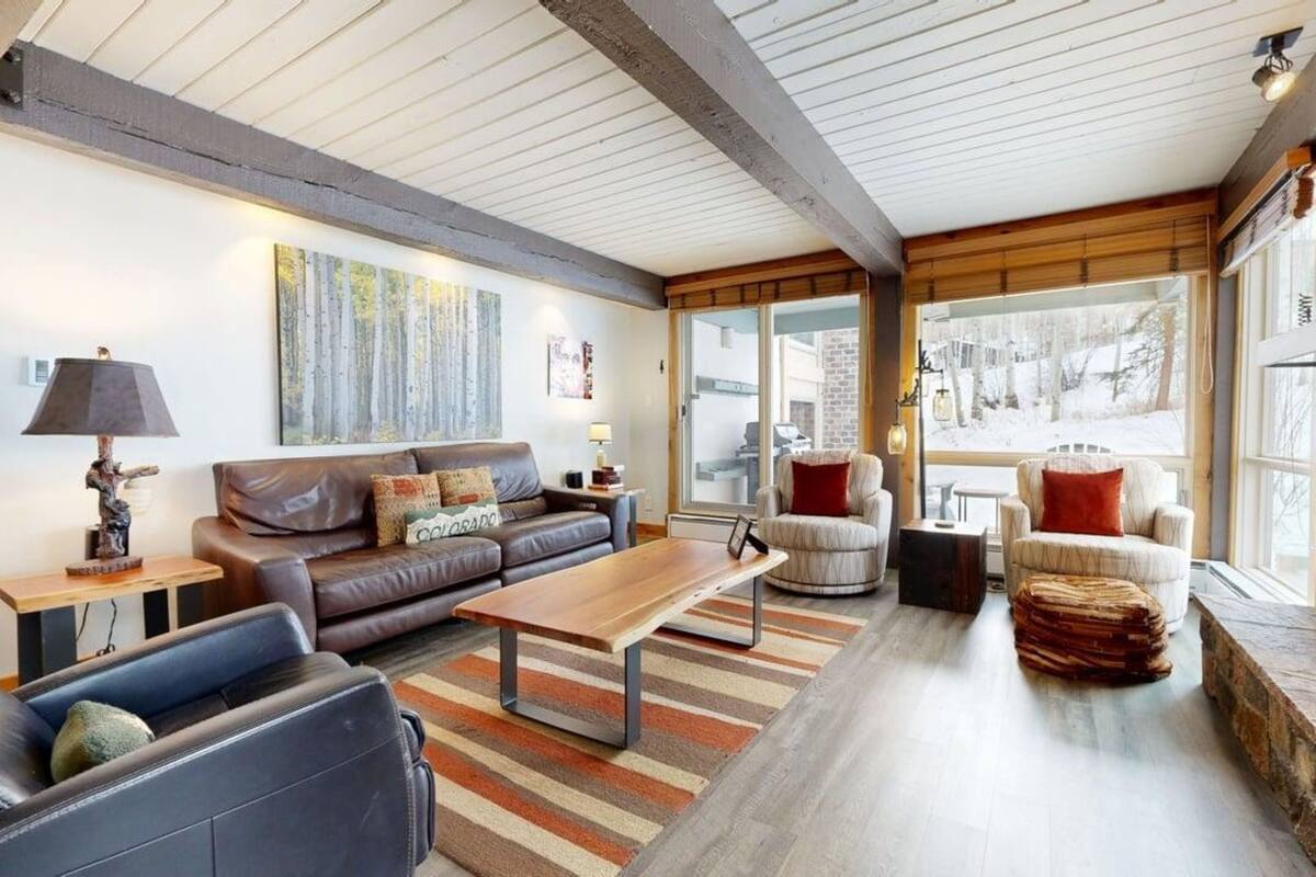 A modern living room with a brown leather sofa, a wooden coffee table, and a striped rug. There are two armchairs and a small table with a lamp. The room has large windows and a view of a snowy landscape.