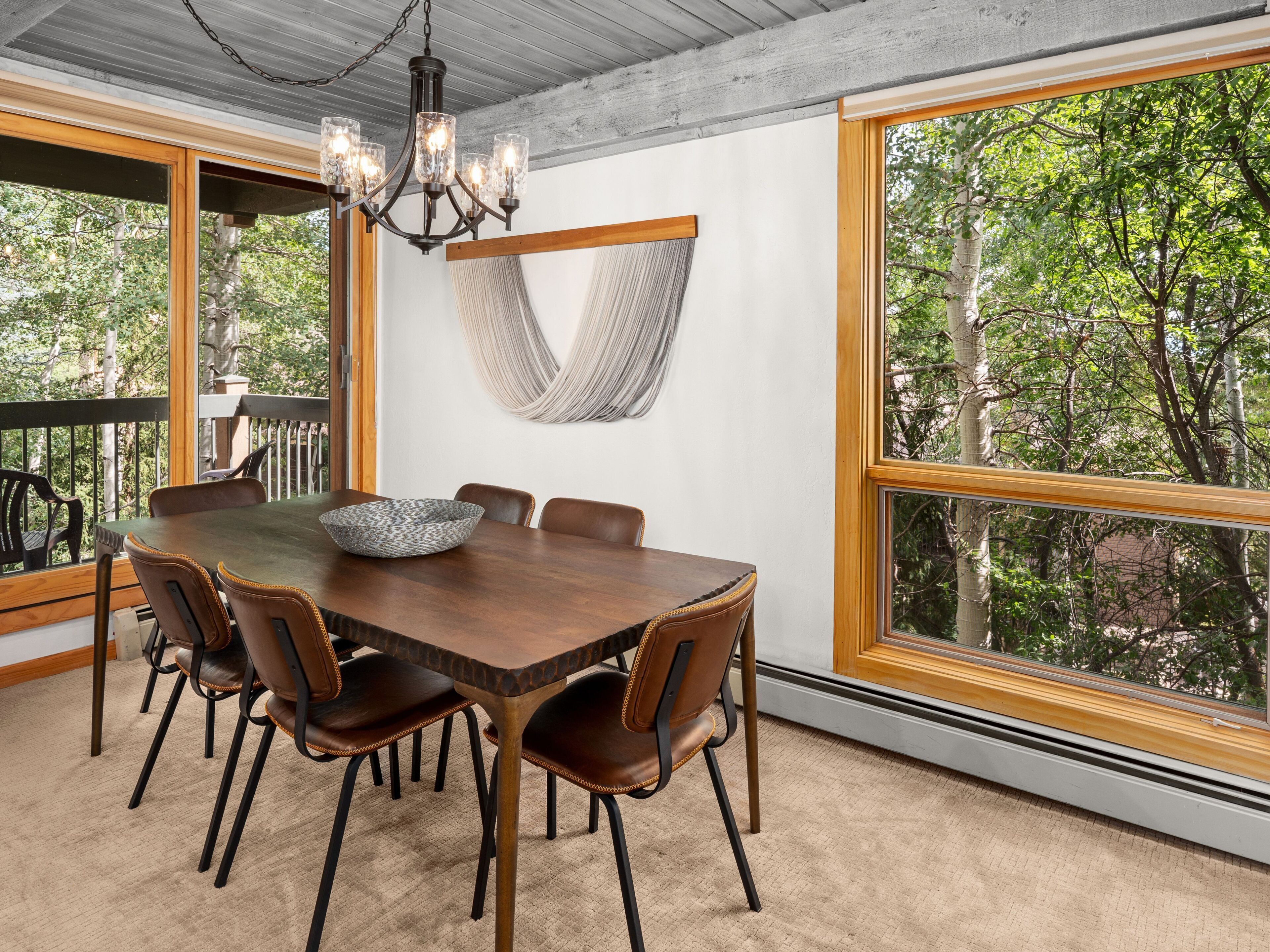 A dining area with a large table, six chairs, a chandelier, and a view of trees through large windows.