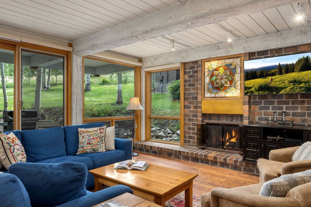 A cozy living room with a fireplace, large windows, and a stone wall.