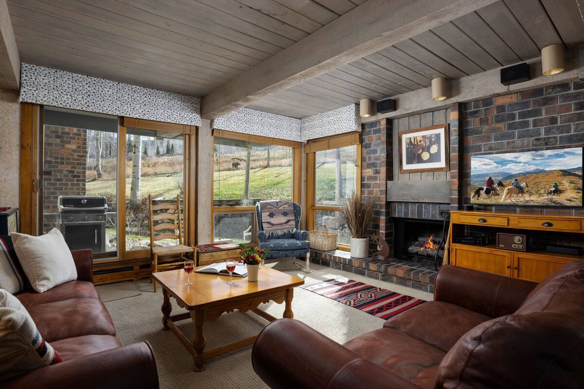 A cozy living room with a fireplace, leather sofas, and a large window overlooking a grassy area.