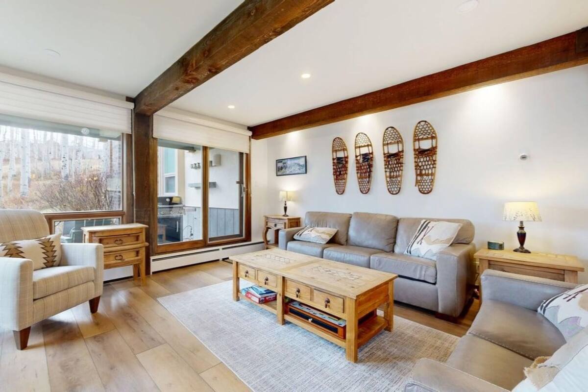 A cozy living room with a wooden coffee table, a grey sofa, and a white armchair. There are two side tables with lamps, and a large window with blinds.